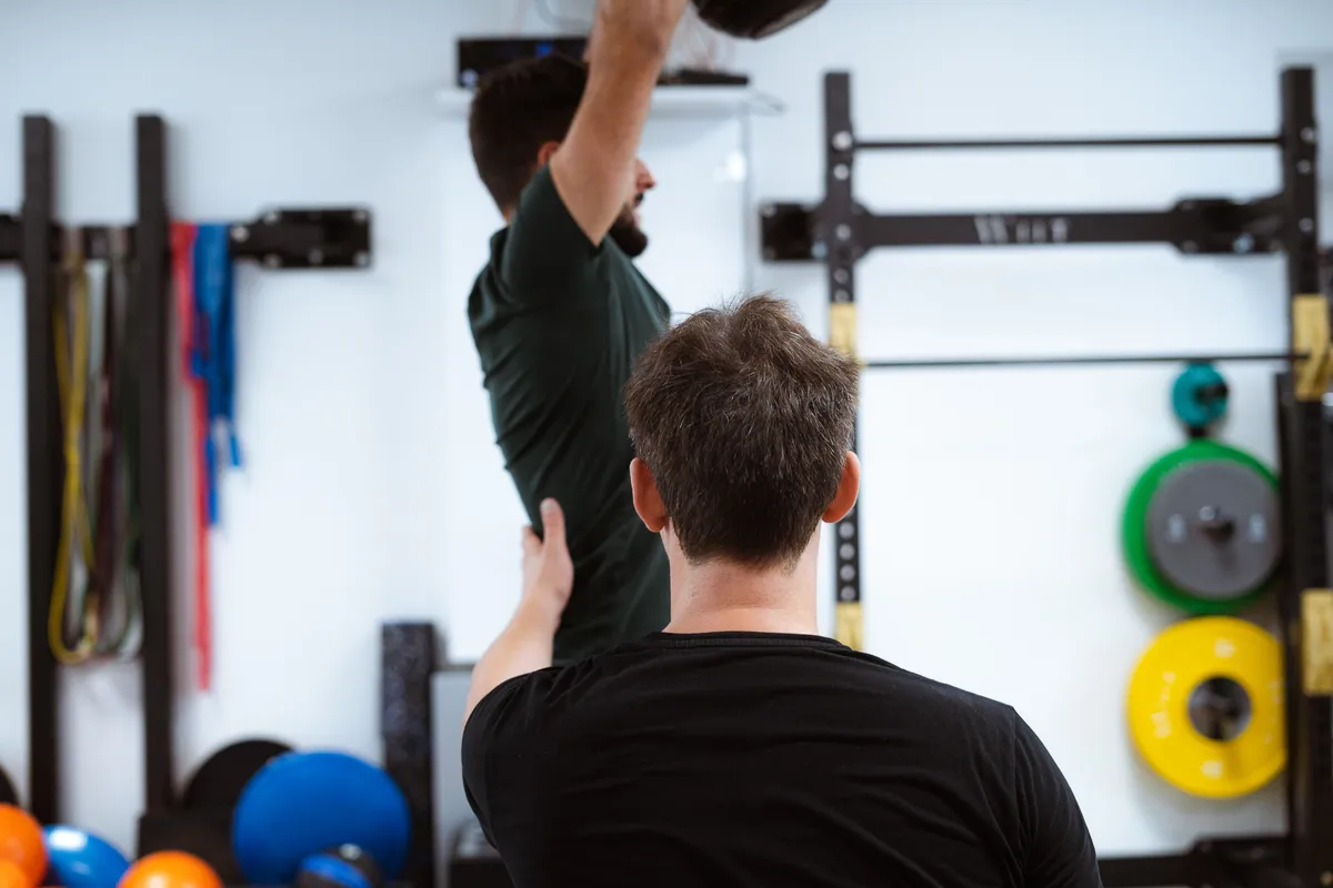 A man demonstrates an overhead movement to another member next to a squat rack