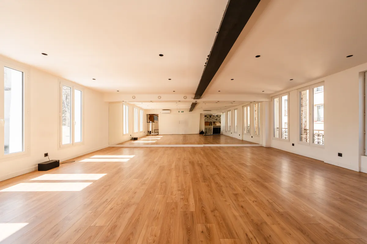 Large empty studio with light wood floor, tall windows, wall mirrors and an exposed steel beam